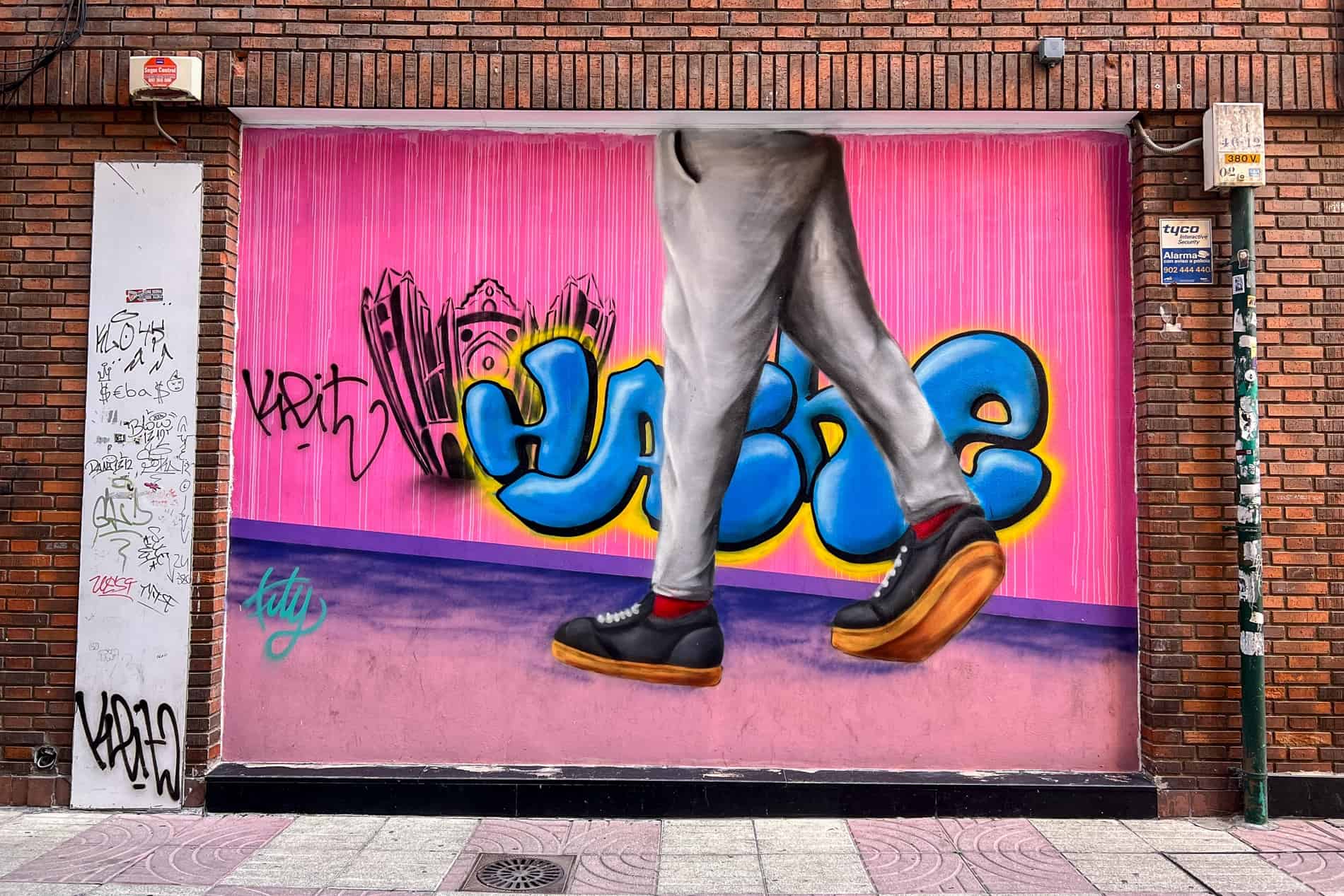 Arte callejero de la mitad inferior de una persona que pasa frente a una pared rosa con letras azules de grafiti. 