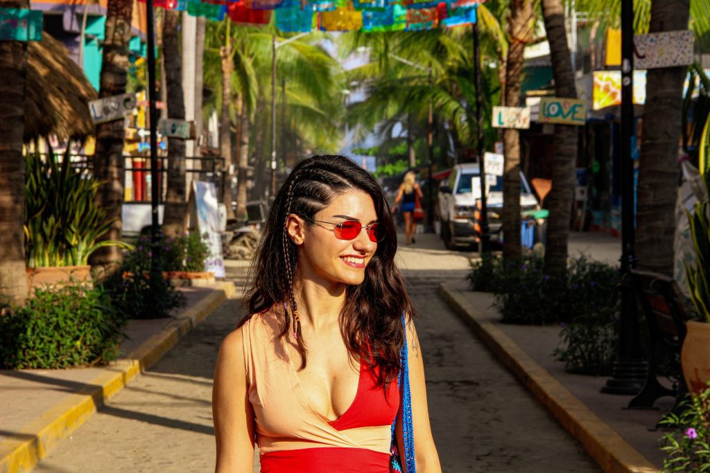 Una mujer con gafas de sol rojas y cabello trenzado camina por una calle con bandera en Sayulita