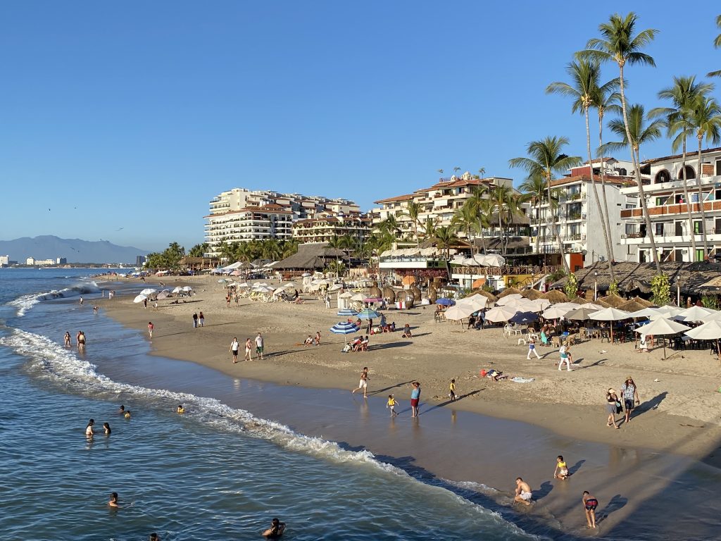 Una playa bordeada de altos edificios de apartamentos blancos y palmeras, mucha gente jugando en el agua.