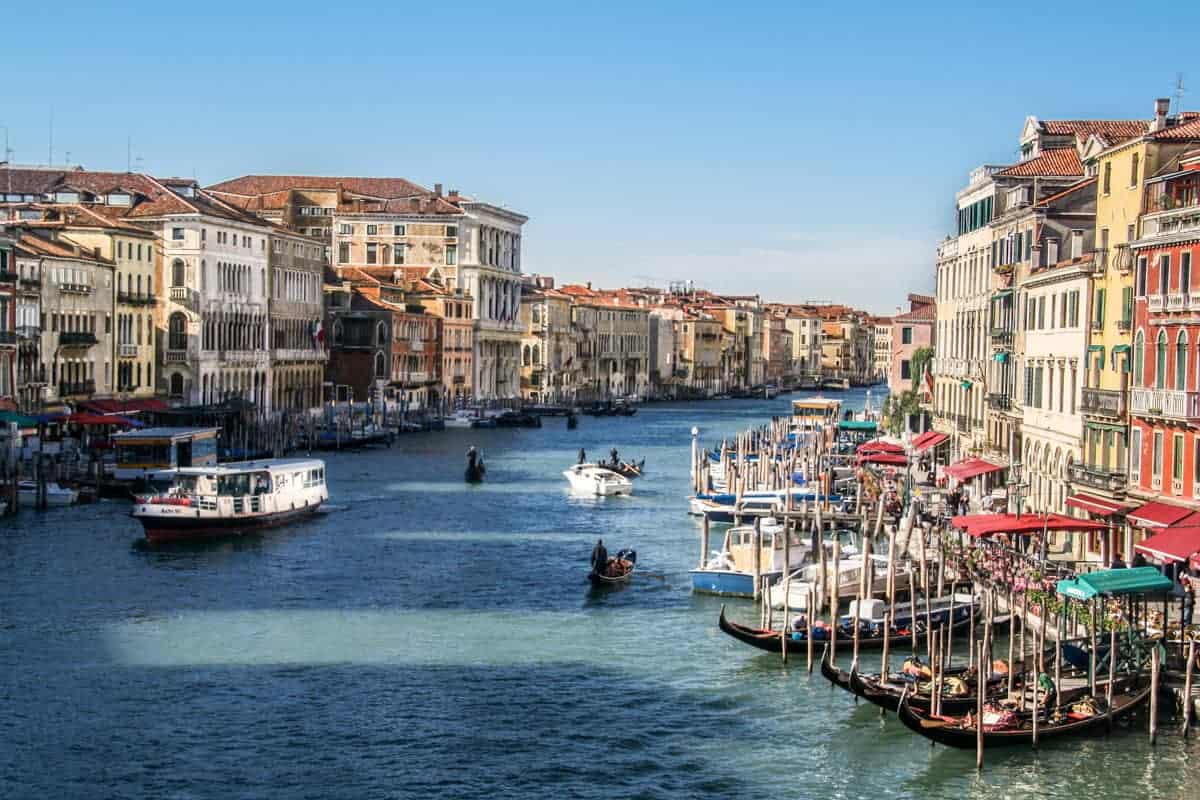 Vista de barcos y góndolas en el gran canal azul profundo de Venecia, flanqueado por edificios en tonos dorados pastel.