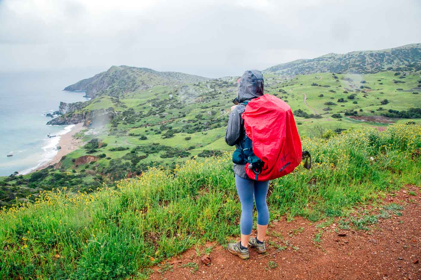 Mujer en un sendero con vistas a la isla y al océano.  El cielo está lluvioso y ella lleva un chubasquero.
