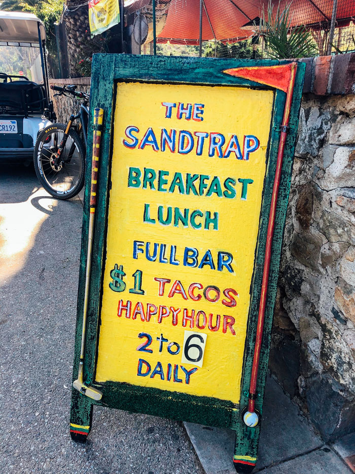 Letrero amarillo pintado que dice "La trampa de arena.  Desayuno, almuerzo, bar completo.  Hora feliz de tacos a $1"